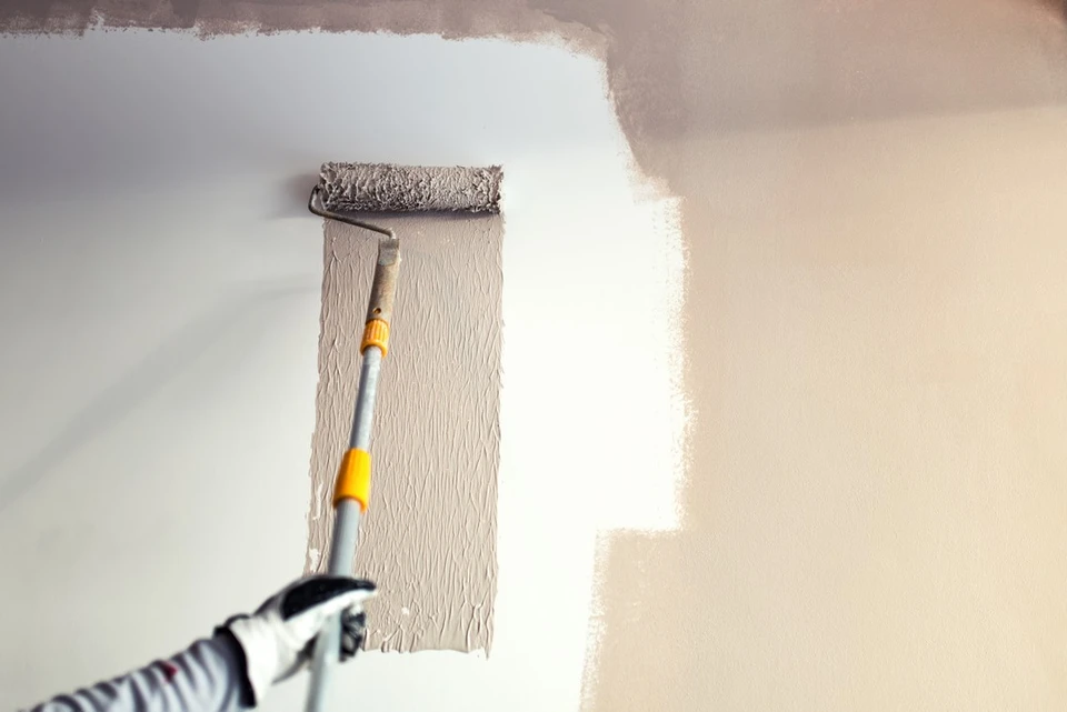 Main entre un mur beige et blanc en cours de peinture avec rouleau à peinture tenu par une personne portant des gants de protection rayés noir et blanc